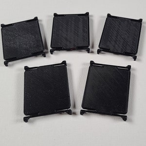 Może przedstawiać: Pięć czarnych, kwadratowych obiektów wydrukowanych w 3D o teksturowanej powierzchni i małych nóżkach. Obiekty są ułożone na białej powierzchni. Każdy ma prostokątny kształt z lekko pochyloną górą i dołem.