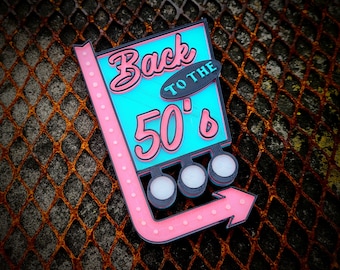 Peut inclure: Une enseigne rétro avec le texte "Back to the 50's". L'enseigne présente un fond turquoise avec des lettres roses et une flèche rose avec des points lumineux blancs. Elle a une bordure noire et trois ampoules blanches.
