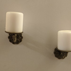May include: Two wall-mounted candle holders with white candles. The candle holders have a decorative, ornate design in a bronze color. The candles are cylindrical and have a smooth, white surface. The background is a light gray wall.