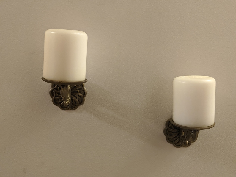 May include: Two wall-mounted candle holders with white candles. The candle holders have a decorative, ornate design in a bronze color. The candles are cylindrical and have a smooth, white surface. The background is a light gray wall.