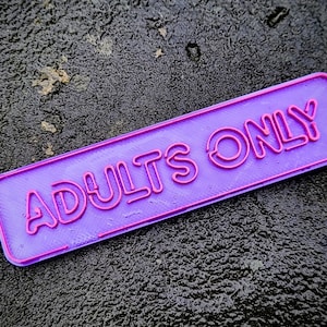 Pode incluir: Uma placa retangular roxa com as palavras "ADULTS ONLY" em letras rosa em relevo. A placa tem uma borda rosa e está sobre um fundo escuro e texturizado.