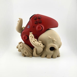 May include: A red video game controller rests on a beige octopus-shaped sculpture. The controller has black buttons and a directional pad. The octopus has detailed tentacles and a textured surface, creating a unique gaming accessory.