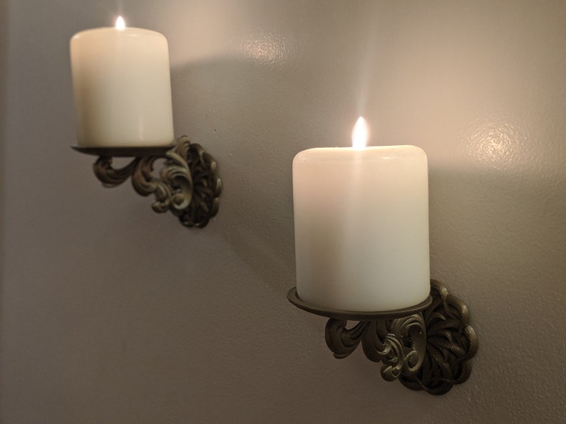 May include: Two lit white pillar candles in ornate, antique-style wall sconces. The sconces are made of a dark metal with a decorative, swirling design. The candles are centered in the sconces, casting a warm glow.