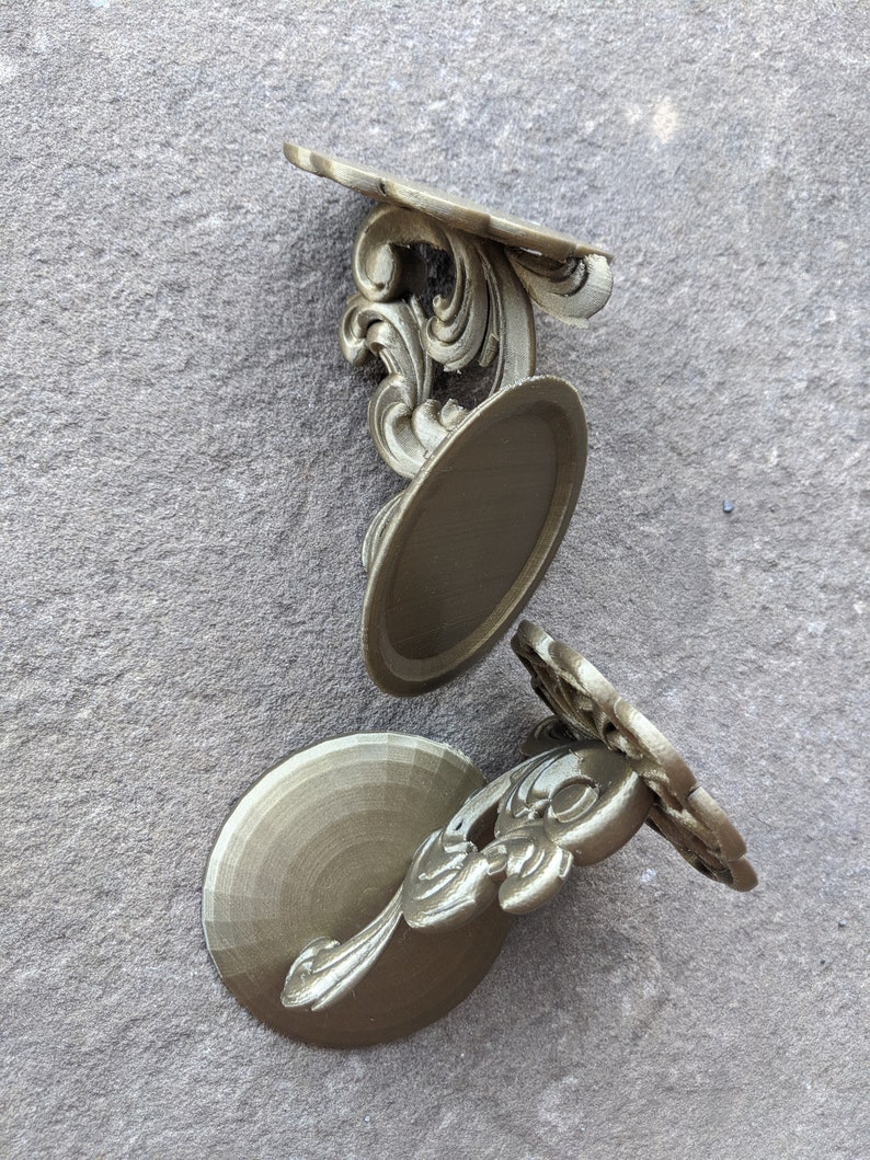 May include: Three ornate, gold-colored decorative wall shelves. Each shelf features a circular base, a decorative floral design, and a curved top. The shelves are arranged on a textured gray surface.