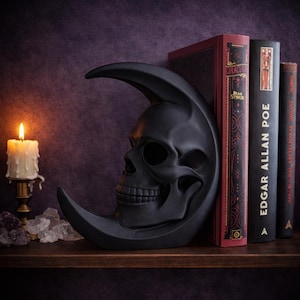 May include: A black skull-shaped bookend in the form of a crescent moon. The bookend is placed on a wooden shelf next to several antique books with titles like "Dracula" and "Edgar Allan Poe". A lit candle and crystals are also on the shelf.