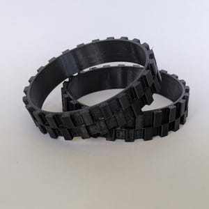 Puede incluir: Tres orugas negras, circulares, impresas en 3D, con un patrón rectangular repetido en el borde exterior. Las orugas están apiladas unas sobre otras. Están hechas de un material negro mate. La oruga más grande está en la parte inferior y la más pequeña en la superior.