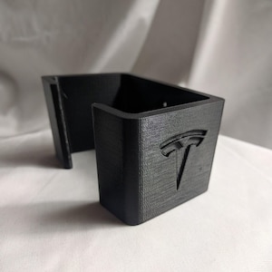 May include: A black, 3D-printed car console organizer with the Tesla logo. The organizer has a rectangular shape with a slot for storage and is designed to fit in a car's console.