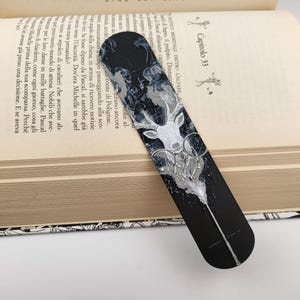 May include: A black bookmark with a white deer design, lying on an open book. The bookmark features a detailed illustration of a deer head with geometric shapes and a flowing, abstract design. The book's pages are visible, with text and the number 353.