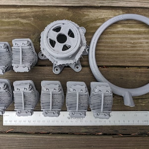 May include: A collection of gray 3D-printed model airplane engine parts, including cylinders, a circular ring, and a central engine component. A ruler shows measurements in inches. The parts are arranged on a wooden surface.