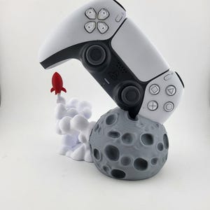May include: A white and black video game controller rests on a gray moon-shaped base with a red rocket taking off. The controller has black buttons and a white body. The moon base has craters.