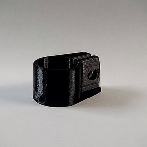 May include: A black 3D-printed object, possibly a clamp or holder, with a cylindrical opening and a rectangular base. The object has a textured surface and appears to be made of plastic. It is set against a plain white background.