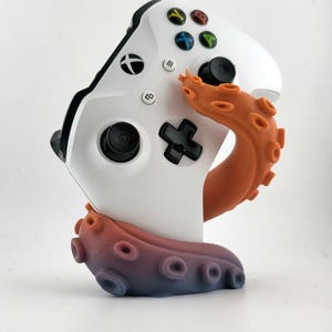 May include: White Xbox controller with colorful buttons, resting on a 3D-printed stand shaped like an orange and purple octopus tentacle. The controller has black joysticks and buttons.