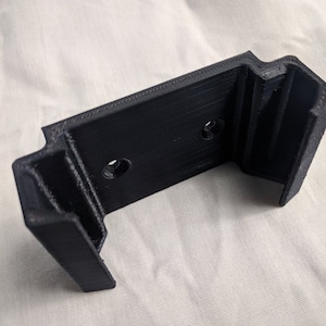 Può includere: Una staffa nera stampata in 3D con una base rettangolare e due fori di montaggio. La staffa ha due bracci angolati che si estendono dalla base, creando una forma a U. L'oggetto è realizzato in materiale nero opaco.