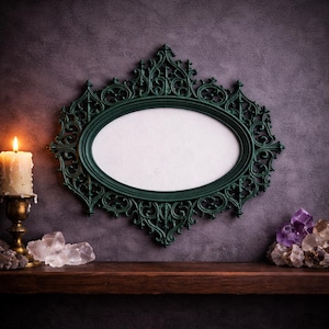 Puede incluir: Un marco ovalado verde oscuro y ornamentado con detalles intrincados cuelga de una pared morada oscura texturizada. Una vela encendida en un soporte dorado y cristales se encuentran en una repisa de madera, creando una estética mística.