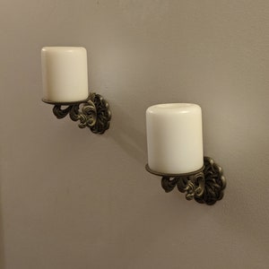 May include: Two wall-mounted candle holders with ivory-colored candles. The holders have an ornate, antique brass finish with a floral design. The candles are cylindrical and sit in a circular base.