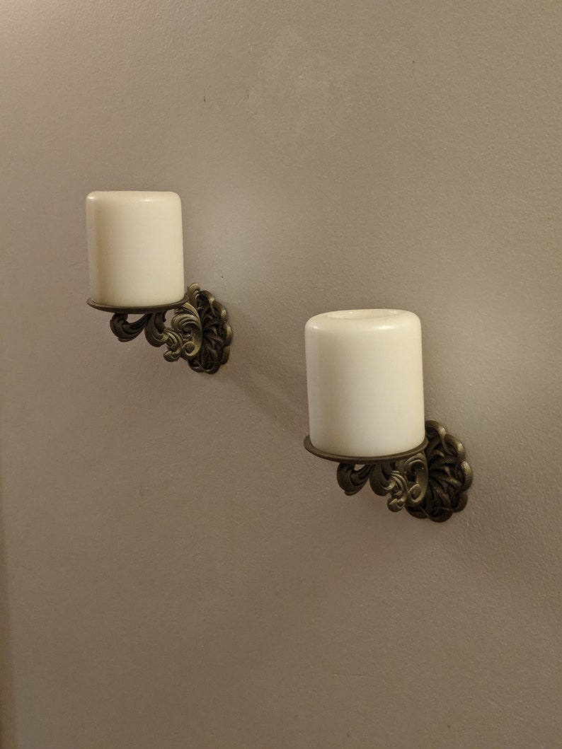 May include: Two wall-mounted candle holders with ivory-colored candles. The holders have an ornate, antique brass finish with a floral design. The candles are cylindrical and sit in a circular base.