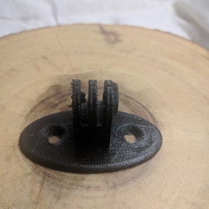May include: A black 3D-printed camera mount with a three-pronged fork design. The mount has an oval base with two holes for screws. The mount is designed to attach to a wooden surface.
