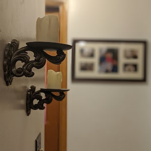 May include: Two ornate, dark metal wall sconces with white candles. The sconces have a decorative, swirling design and are mounted on a white wall. A framed picture is visible in the background.