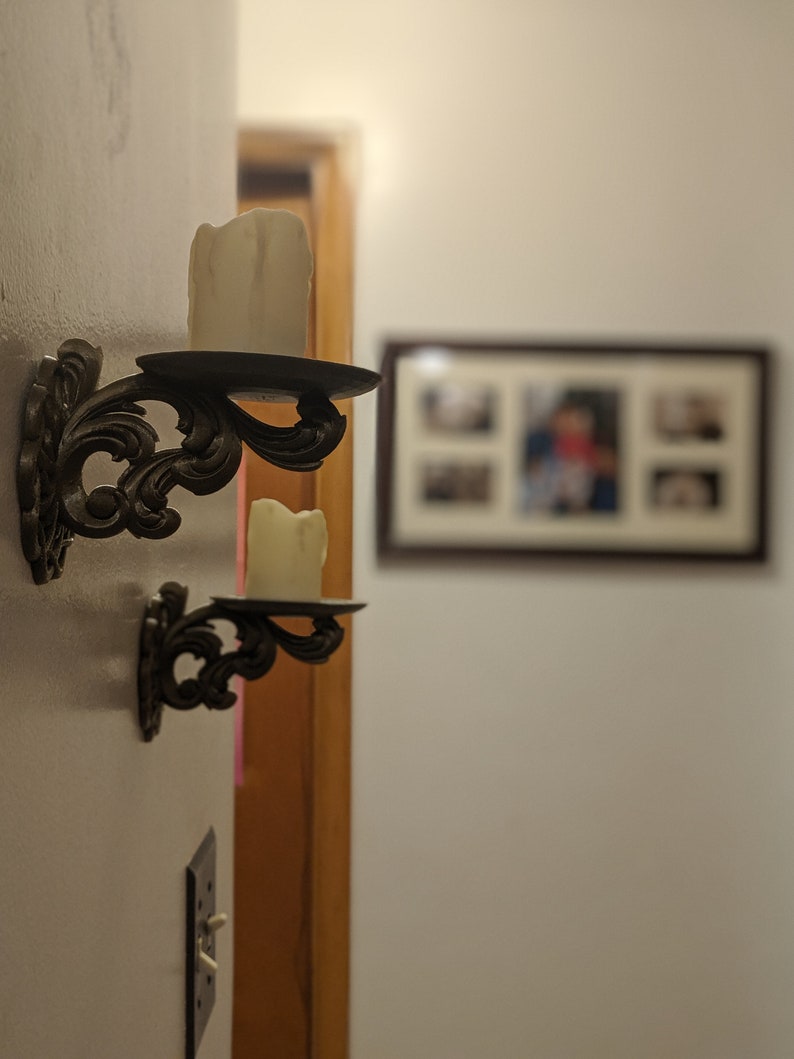 May include: Two ornate, dark metal wall sconces with white candles. The sconces have a decorative, swirling design and are mounted on a white wall. A framed picture is visible in the background.