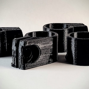 May include: Four black 3D-printed plastic components. Each has a cylindrical cup shape with a rectangular base and a hole. The objects are arranged on a white surface, with the lighting creating shadows and highlighting the textures of the plastic.