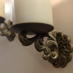 May include: Ornate, gold-colored wall sconces with a decorative, swirling design. Each sconce holds a white candle. The sconces are mounted on a light-colored wall, creating a classic and elegant look. Perfect for home decor.