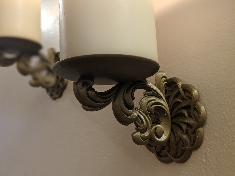 May include: Ornate, gold-colored wall sconces with a decorative, swirling design. Each sconce holds a white candle. The sconces are mounted on a light-colored wall, creating a classic and elegant look. Perfect for home decor.