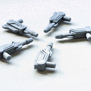 May include: Five gray, 3D-printed, futuristic-looking components are arranged on a white surface. Each component has a rectangular body with geometric details and a cylindrical extension. The components appear to be small mechanical parts.