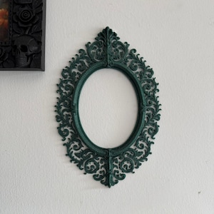 Puede incluir: Un marco de fotos ovalado verde oscuro con intrincados adornos. El marco está vacío y montado en una pared blanca. El marco tiene una parte superior puntiaguda y una base decorativa. Un marco negro con detalles de calaveras y rosas es visible en la esquina superior izquierda.