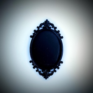 May include: A black, oval picture frame with elaborate scrollwork. The frame has a matte finish and is set against a white background. The frame's design is symmetrical, with decorative elements at the top and bottom.