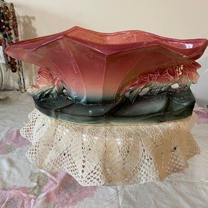 May include: A large, pink and green ceramic planter shaped like a flower with a scalloped rim. The planter is sitting on a white crocheted doily.
