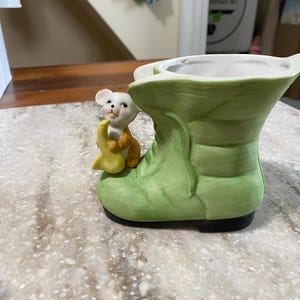 May include: A ceramic planter shaped like a green boot with a small mouse holding a yellow crescent moon. The boot has a black sole and a white interior. The mouse is brown and white.