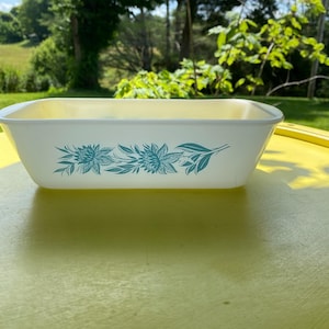May include: A rectangular off-white glass baking dish featuring a horizontal band of teal floral patterns along the center of the exterior. The dish has integrated handles on both ends and a smooth, glossy finish throughout the entire vessel.