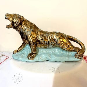 May include: A gold-colored ceramic tiger figurine with an open mouth, resting on a light blue base. The tiger has a textured surface and is in a roaring pose. The figurine is a decorative item.