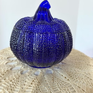 May include: A cobalt blue glass pumpkin-shaped container with a textured, pebbled surface and a smooth, curved stem. The object features a horizontal seam separating the lid from the base, showcasing a vibrant, translucent deep blue color.