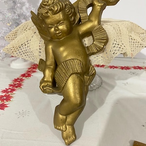 May include: A golden cherub figurine with detailed wings and a flowing garment. The cherub is posed in a dynamic stance, holding a decorative element. The figurine is likely made of a metallic material and is a decorative item.