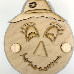 May include: A wooden cutout of a scarecrow face with a hat, eyes, nose, and stitched mouth. The cutout is unfinished and ready to be painted or decorated.