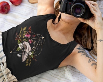 May include: A woman wearing a black tank top with a floral and skull design. The tank top has a geometric pattern and a skull with flowers and leaves. The woman is lying on a white blanket with a picnic basket and food in the background.