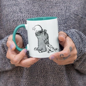 Puede incluir: Una taza de cerámica blanca con un borde y un asa de color turquesa. La taza presenta una ilustración en blanco y negro de una criatura con una estrella en la cabeza, sosteniendo una manta con estrellas.