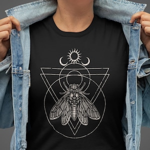 May include: A black t-shirt with a white graphic design of a moth inside a geometric shape with a sun and moon above it.