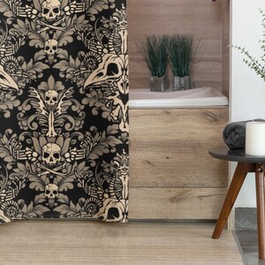 Op de afbeelding: Een zwart en gouden douchegordijn met een schedel en gekruiste beenderen patroon. Het gordijn hangt in een badkamer met een houten badkuip en een klein bijzettafeltje met een kaars en een vaas met groen.