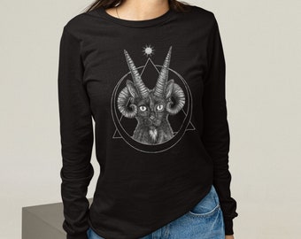 Pode incluir: Uma camiseta preta de manga comprida com um gráfico de um gato preto com chifres e chifres de carneiro em um círculo com um triângulo e uma estrela. A modelo está vestindo jeans azul e um chapéu de pescador preto.