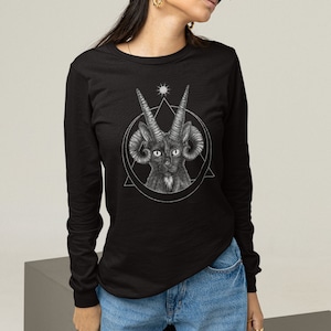 Pode incluir: Uma camiseta preta de manga comprida com um gráfico de um gato preto com chifres e chifres de carneiro em um círculo com um triângulo e uma estrela. A modelo está vestindo jeans azul e um chapéu de pescador preto.