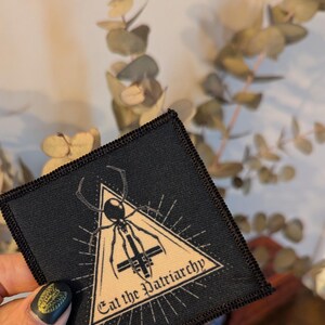 May include: Black iron-on patch featuring a black spider inside a beige triangle with the text "Eat the Patriarchy" in beige lettering.