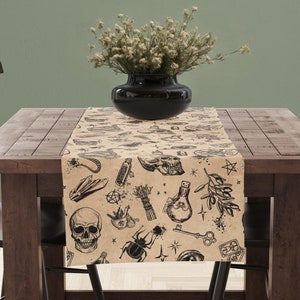 May include: A brown table runner featuring black illustrations of skulls, crystals, keys, and potion bottles. The runner is on a wooden table, with a black vase and dried flowers.