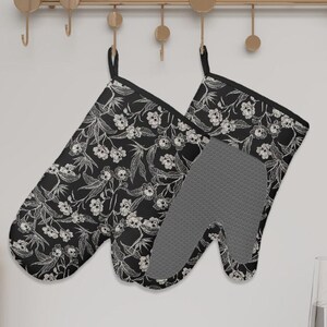 May include: A pair of black oven mitts with a white floral pattern, hanging on a gold-colored rack. The mitts have a black trim and a gray, textured interior. Kitchenware.