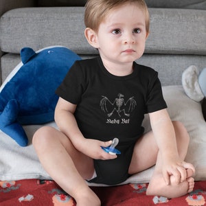 May include: A baby wearing a black bodysuit with a white skeleton bat graphic and the text "Baby Bat". The baby is sitting on a red blanket with a pattern of white goats.