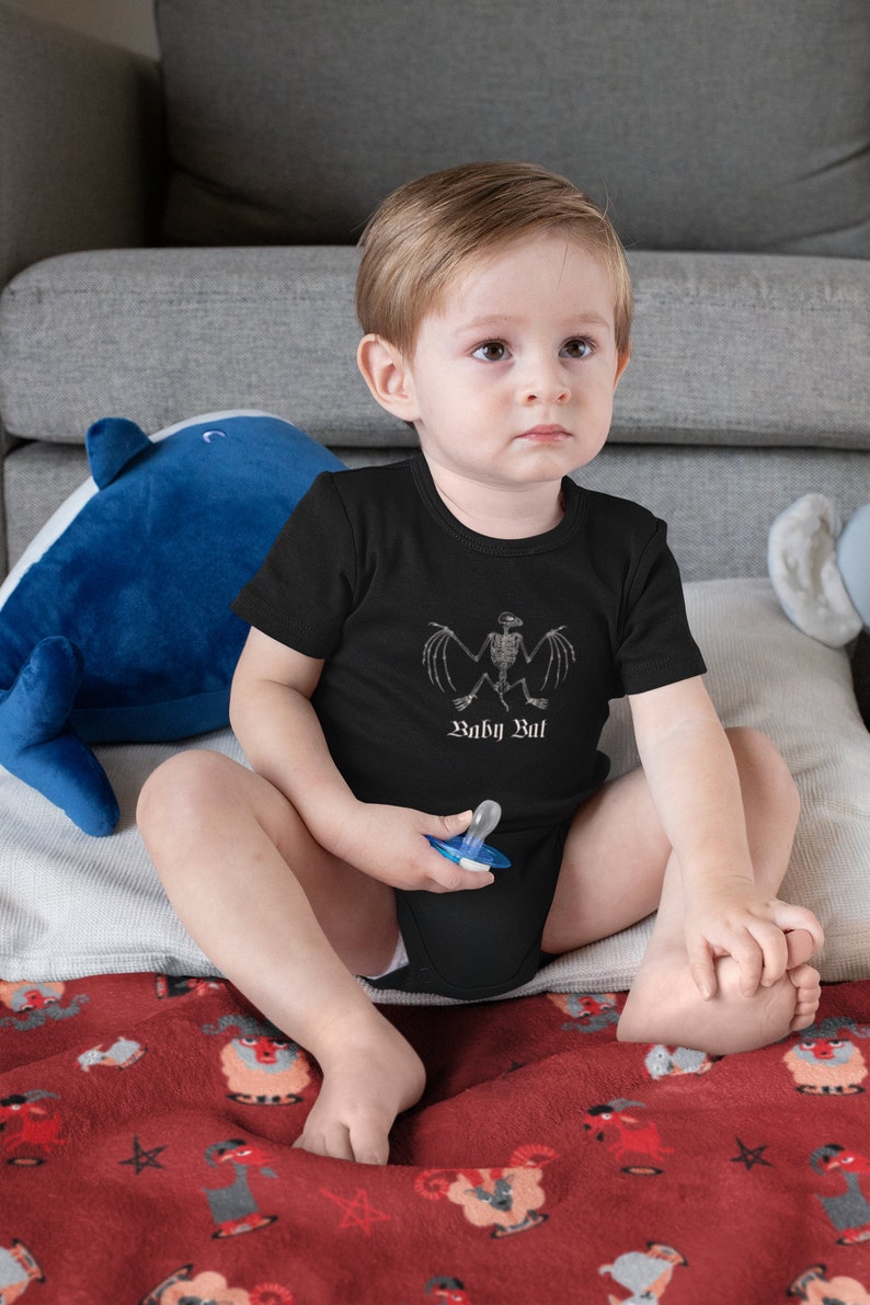 May include: A baby wearing a black bodysuit with a white skeleton bat graphic and the text "Baby Bat". The baby is sitting on a red blanket with a pattern of white goats.