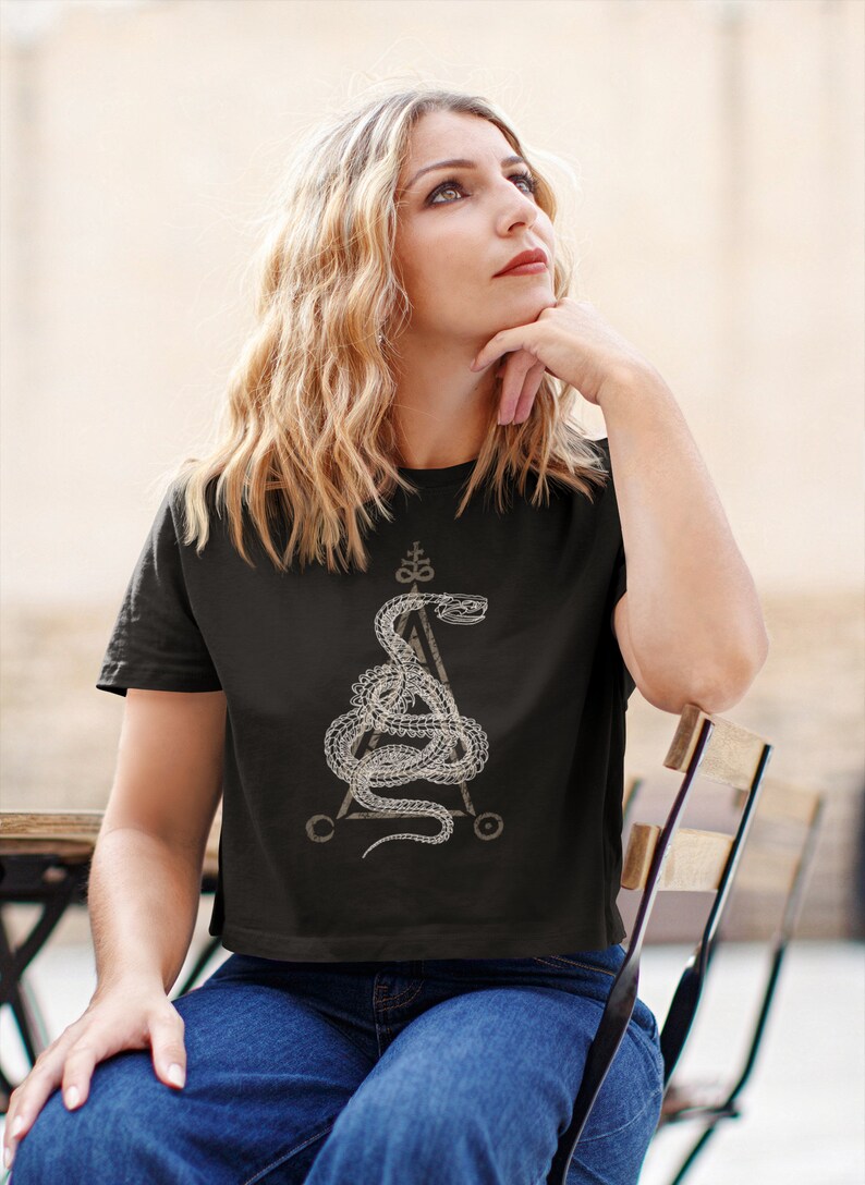 May include: A black cropped t-shirt with a white graphic of a snake wrapped around a triangle with a symbol at the top. The snake is stylized and has a detailed design.
