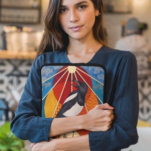 May include: A woman is holding a colorful laptop sleeve with a black and white magpie illustration. The sleeve has a geometric design with a sun, clouds, and a rainbow.