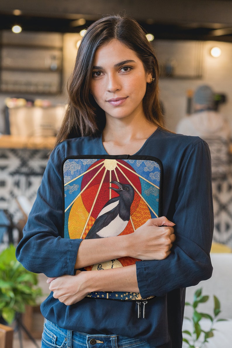 May include: A woman is holding a colorful laptop sleeve with a black and white magpie illustration. The sleeve has a geometric design with a sun, clouds, and a rainbow.